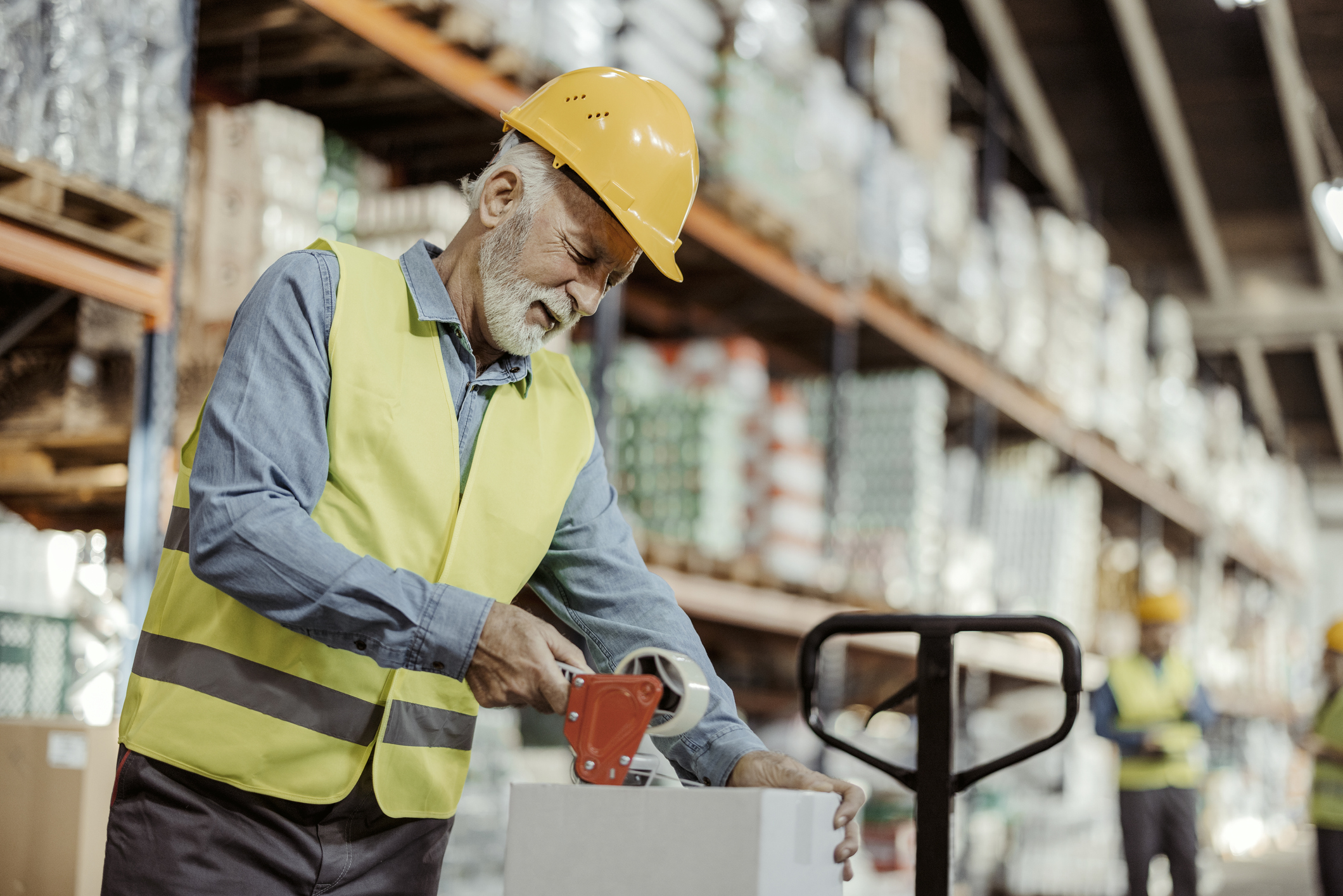 Senior male worker packing cardboard box with tape gun dispenser in warehouse