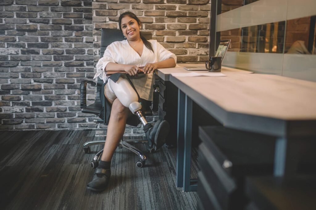 A woman with a prosthetic leg sits smiling on an office chair beside a desk with a tablet and cup. She is wearing a white blouse and black pants.