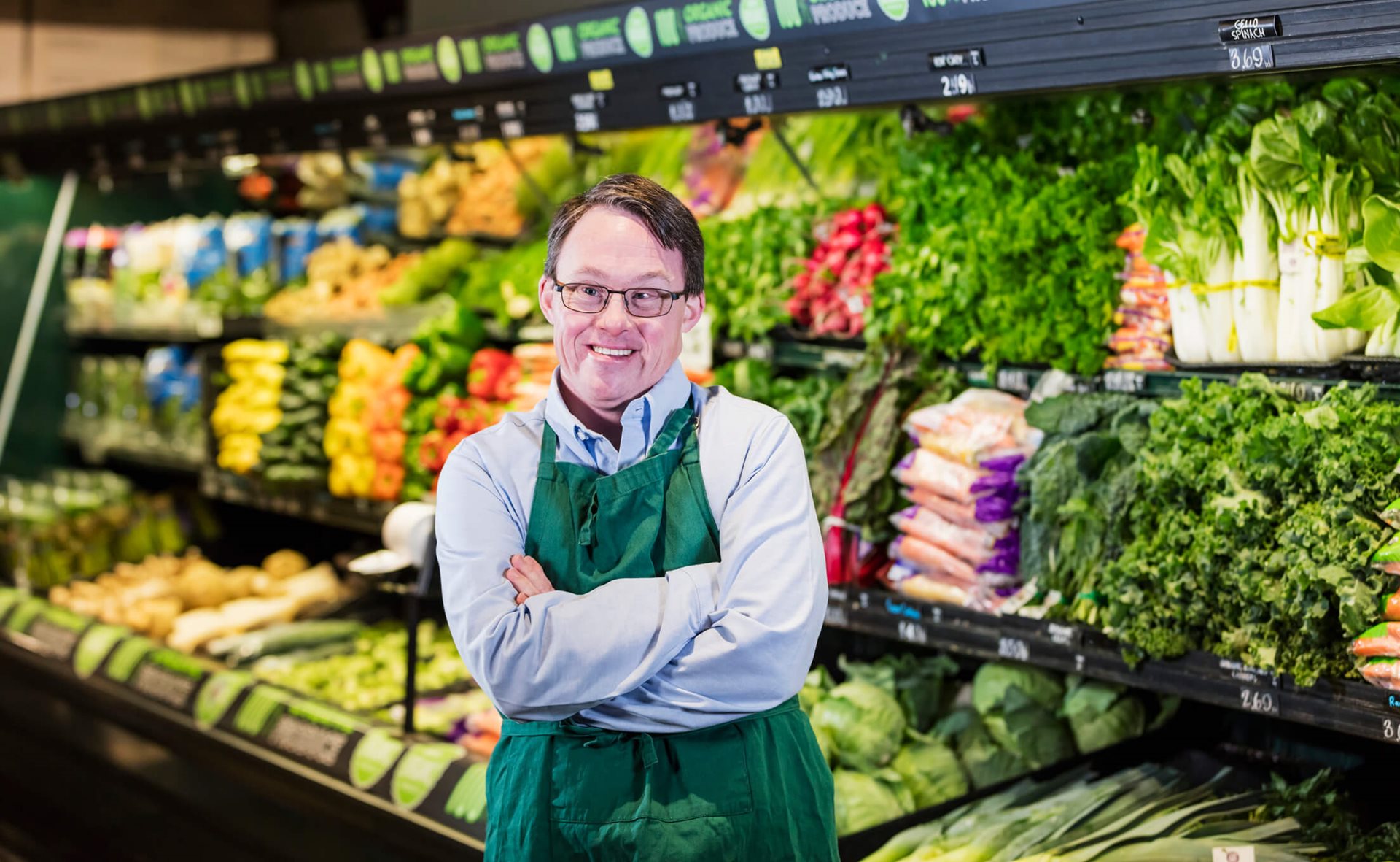 A man with a disability wearing a green apron stands in front of a colorful produce section, showcasing fresh fruits and vegetables.