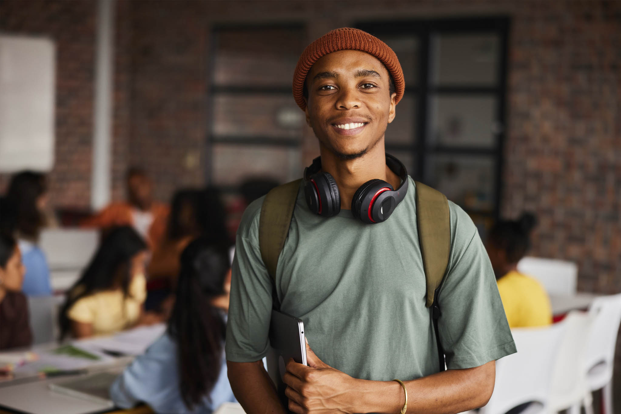 Portrait of a young male college student wearing headphones and a beanie smiling while standing in a classroom with students behind him