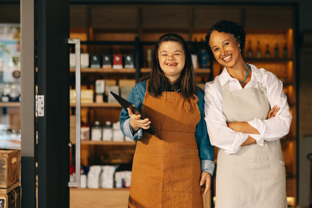 A successful shop owner smiling warmly at the camera alongside her employee, who has a visible disability, both exuding confidence and positivity in their workplace environment.